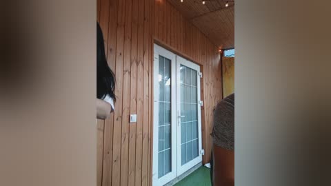 A video of a cozy, rustic indoor space featuring wooden walls, a white-framed glass door, and a green carpet. A person in a black shirt stands near the door, and there are string lights and a wooden bench in the background.