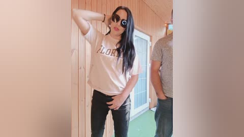 Video of a slender, light-skinned woman with long black hair, wearing a white t-shirt with \"LOVE\" and black pants, posing confidently in a sunlit hallway.