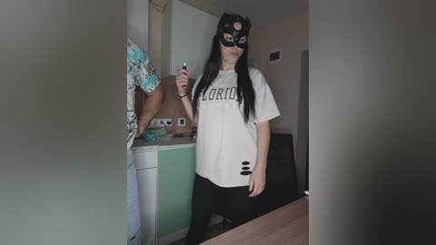 Video of a young woman with long black hair, wearing a black mask and white T-shirt with \"LORI\" printed in black, taking a selfie in a dimly lit room with green and white walls.