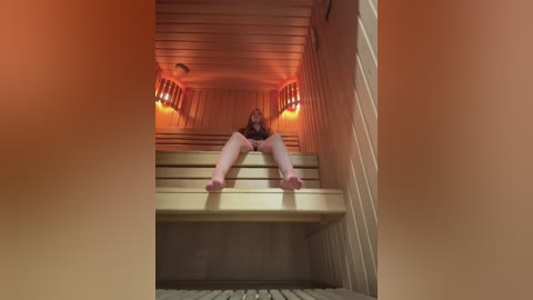 Video of a woman with long brown hair, sitting on a wooden sauna bench, legs spread, wearing a black top, in a dimly lit sauna with orange lighting.