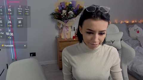 Video of a woman with short dark hair, wearing a beige ribbed sweater, sitting in a modern living room with a floral arrangement, digital clock, and white chair.