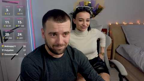 Video of a man with short dark hair, wearing a dark shirt, sitting in a modern room with a woman in a white turtleneck and black shorts.
