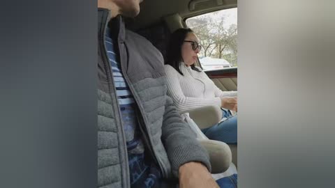 A video of a man and woman sitting in a car, both wearing casual attire, the woman with dark hair and glasses, and the man with a jacket. The car's interior is visible, and the background shows trees and a cloudy sky.