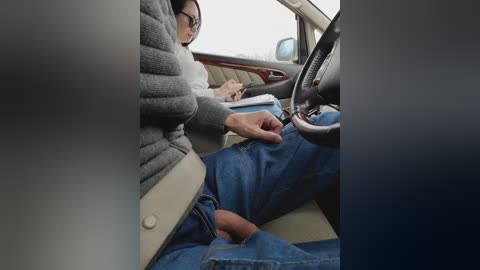Video of a woman in a gray sweater and glasses, sitting in a car, buckling her seatbelt, while another person, possibly a driver, is focused on paperwork in the background.