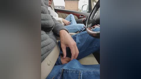 Video of a person in a car, wearing a gray sweater and blue jeans. They are adjusting the seatbelt, visible in the background is the car's interior and the steering wheel.