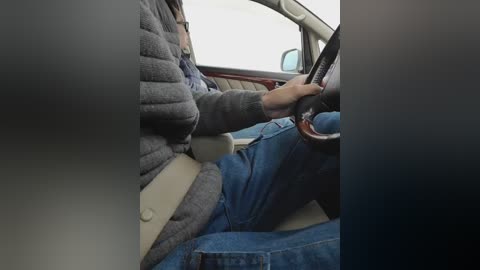 Video of a person wearing a grey sweater and blue jeans, driving a vintage car with beige leather seats, steering wheel, and dashboard.