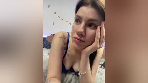 Video of a young woman with light skin, long brown hair, and blue bra, leaning on her elbows, looking thoughtful. Background features a bed with grey sheets, string lights, and a blurred, nude male figure.