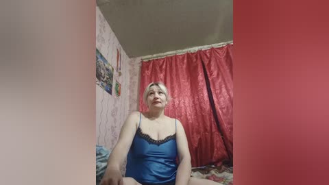 Video of a plus-sized, fair-skinned woman with short blonde hair, wearing a blue camisole with black lace trim, sitting on a bed with red curtains and floral wallpaper in the background.