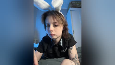 Video of a young person with pale skin and short brown hair, wearing a white bunny ears headband, black shirt, and gray blanket, indoors with blue lighting.