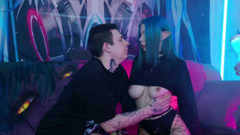 A video of a couple, a man with short brown hair and a woman with blue hair, both in black clothing, kissing passionately in a dimly lit room with neon lights and abstract wall art.