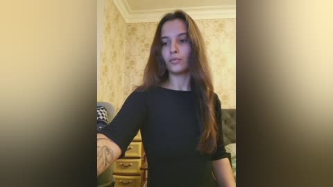Video of a young woman with long brown hair, wearing a black top, standing in a room with patterned wallpaper, a dresser, and a chair.