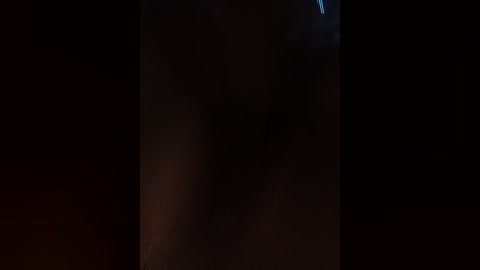 A dimly lit video of a person's face with a blurred, indistinct shadowy background. The image is grainy, suggesting low light or a night setting.