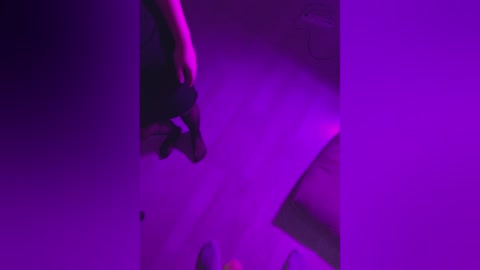 A dimly lit video in purple hues captures a person, possibly female, wearing a black shirt and holding a phone, standing in an abstract, shadowy setting.