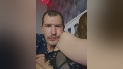 Video of a Caucasian man with short brown hair and a mustache, wearing a blue and white plaid shirt, kissing a woman with long brown hair. The background features a dimly lit room with red string lights.