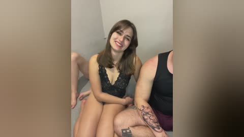 Video of a smiling, slender young woman with shoulder-length brown hair in a black lace bodysuit, sitting on a gray sofa in a minimalist room, partially obscured by two large, dark, rectangular objects.