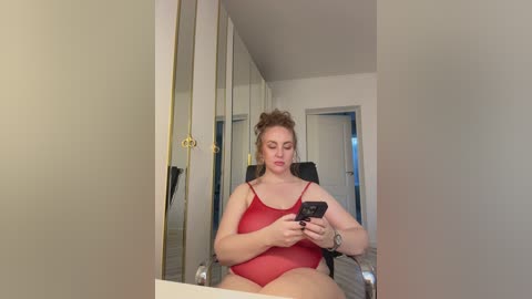 Video of a plus-size woman with fair skin and red hair in a red, sheer, spaghetti-strap bodysuit, seated in a dimly lit room, holding a smartphone.