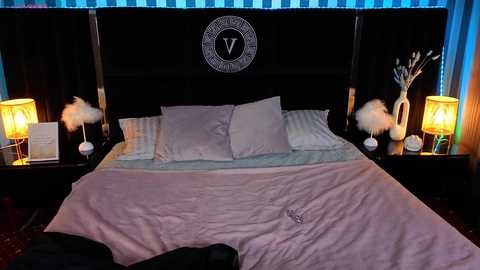 Video of a luxurious, dark-wood bed with white pillows, a pale pink duvet, and a \"V\" emblem on the headboard. Nightstands hold lit lamps, white feather dusters, and blue patterned curtains frame the bed.