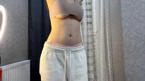 Video of a light-skinned woman in a beige bra and white sweatpants, standing in a dimly lit room with gray-patterned wallpaper and a white lace curtain.