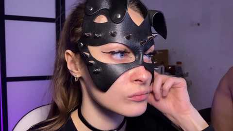 Video of a woman with light skin, brown hair, and blue eyes, wearing a black, spiked leather masquerade mask, with one hand touching her chin. She is dressed in a black outfit, set in a dimly lit room with a purple glow.