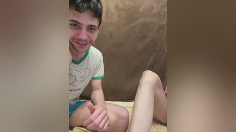 A video of a young man with short dark hair, light skin, and a slim build, wearing a white T-shirt and blue shorts, sitting on a beige blanket, smiling.