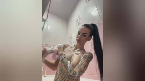 A video of a slender, tattooed woman with long black hair in a ponytail, soaping up in a pink-tiled shower. She has a pale complexion and is wearing a low-cut top.