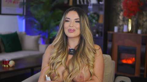 Video of a topless, blonde, fair-skinned woman with long, wavy hair, wearing a black choker, sitting on a beige couch in a cozy, dimly lit living room with green cushions, a fireplace, and potted plants.