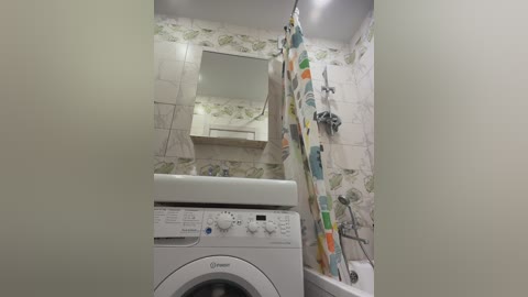 Video of a small, clean bathroom with a white front-loading washing machine, a colorful shower curtain, white-tiled walls, a sink, and a small mirror above it.