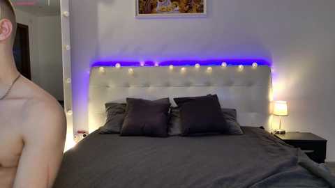 Video of a modern, dimly-lit bedroom with a white tufted headboard, two pillows, and a gray bedspread. A man, topless, stands on the left.