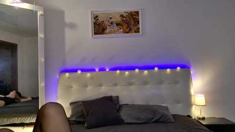 Video of a dimly lit bedroom with a tufted white headboard, gray pillows, a glowing blue LED strip, and a framed painting of a family on the wall.