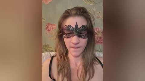 Video of a fair-skinned woman with wavy, light brown hair, wearing a black lace masquerade mask with intricate floral patterns. She has a neutral expression and is dressed in a black camisole. Background features pastel-colored floral wallpaper.