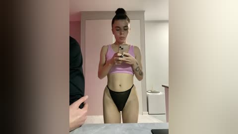 Video of a light-skinned woman with a high bun, wearing a pink crop top and black thong, taking a mirror selfie in a bathroom.
