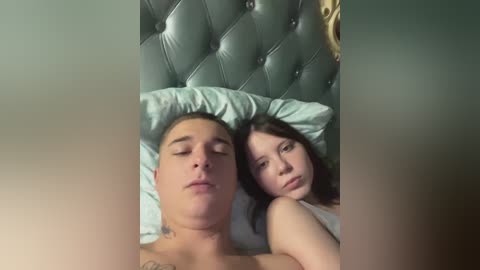 A video shows a sleeping, shirtless young man with a light complexion and a tattoo on his chest, and a woman with long dark hair, both lying on a bed with light blue pillows and a green, tufted headboard.