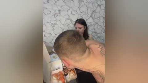 Video of a shirtless man with a shaved head and tattoos, kissing a woman with long brown hair, standing in a room with a white and gray patterned wallpaper.