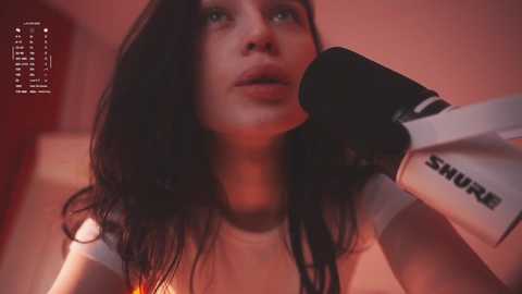 A video of a young woman with long black hair, wearing a white tank top, passionately singing into a SMURF microphone. The background is blurred, with warm, reddish lighting.
