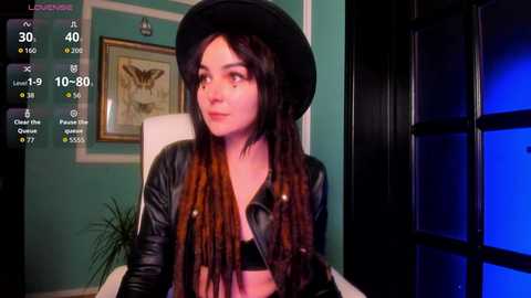 A video of a young woman with long, dark brown dreadlocks, wearing a black leather jacket and hat, sitting in a teal room with a butterfly painting on the wall.