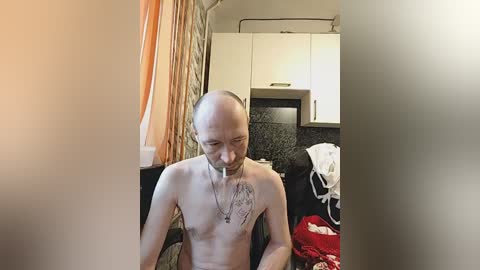 A video shows a thin, bald, fair-skinned man with a cigarette in his mouth, sitting shirtless in a dimly lit, cluttered kitchen with white cabinets and a granite countertop.