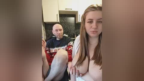 Video of a man and woman in a kitchen. The man, bald with a beard, wears a red and white sweater, sitting on a stool. The woman, with long brown hair, has her breasts exposed, sitting on the counter.