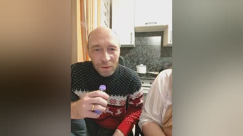 Video of a bald Caucasian man in a black and red Nordic sweater, holding a purple bottle, seated in a modern kitchen with white cabinets and granite countertops.