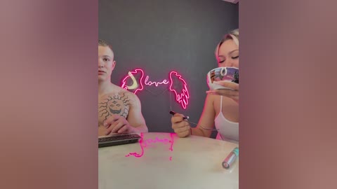 Video of a tattooed, shirtless man and a blonde woman with a phone, both holding a neon sign reading \"Love\" against a dark wall.