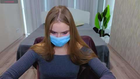 Video of a young Caucasian woman with long, straight light brown hair, wearing a blue surgical mask, gray sweater, and dark brown chair, indoors with light wooden floors, gray curtains, and green plant.