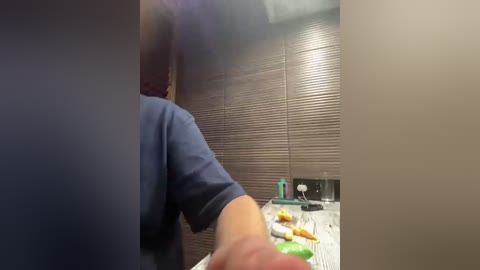 Video of a person in a blue shirt holding a white plate with food, facing a brown-paneled wall and a cluttered table with a green bottle.