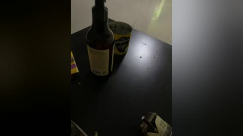 A dimly lit video of a cluttered black table with a dark bottle of wine, a yellow and black can, and scattered papers. The background is blurred, suggesting an indoor setting.