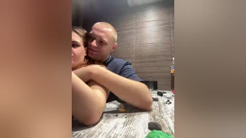 Video of a bald man and a topless woman with blonde hair, both smiling, sitting closely on a bed with a beige blanket, in a dimly lit room with closed blinds.