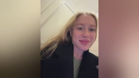 Video of a young blonde woman with fair skin, wearing a black coat, smiling, in a narrow, dimly lit corridor with beige walls and a blurred, warm-toned background.