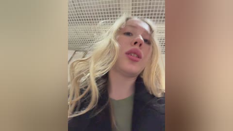 A video of a blonde woman with fair skin and pink lips, wearing a green top and a black jacket, captured from a low angle, with a tiled ceiling background.