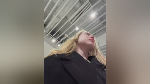 A video of a blonde woman with long hair, wearing a black coat, looking upwards, in a modern, white-walled room with exposed ceiling beams and industrial lighting.