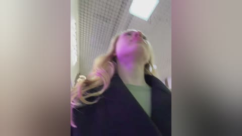 Video of a blonde woman with long hair, wearing a dark jacket, standing in a brightly lit room with a white ceiling and fluorescent lighting.