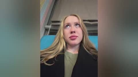 Video of a young Caucasian woman with long, blonde hair and fair skin, wearing a green shirt under a black coat, looking upwards in a public space with blue and beige seating.