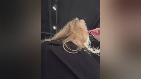A video shows a woman with long blonde hair being styled in a salon. She wears a black jacket. The background is dimly lit with soft lighting.