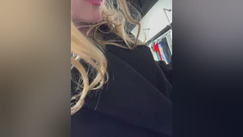 Video of a blonde woman with messy, disheveled hair, wearing a black coat, standing in a cluttered, dimly lit room with hanging clothes and a white wall.